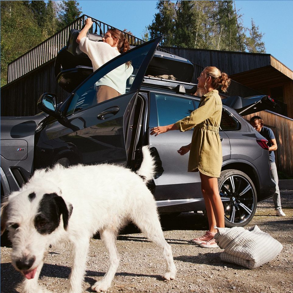 Chien devant femme et homme en train de faire les bagages, BMW en vue latérale en été Révision des vacances BMW – chien devant femme et homme en train de faire les bagages, BMW en vue latérale en été