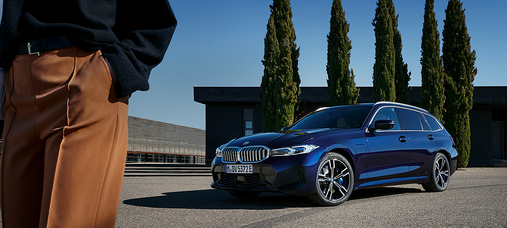 Vista anteriore a tre quarti della BMW Serie 3 Touring LCI G21 Tansanit Blue ferma davanti ad alberi