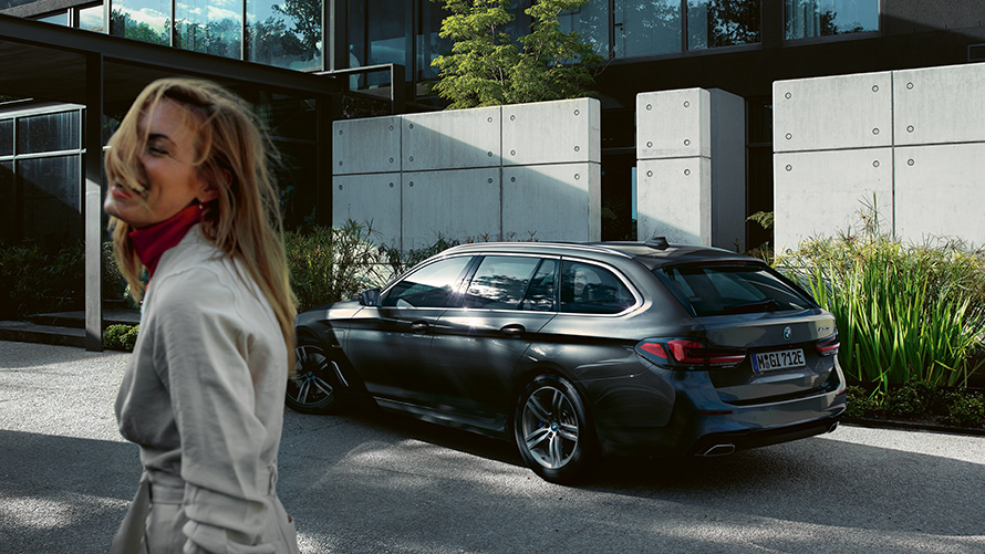 Donna davanti alla BMW Serie 5 Touring G31 LCI Sophisto Grey Brilliant Effect metallizzato vista laterale a 7/8 Donna davanti alla BMW Serie 5 Touring G31 Facelift 2020 Sophisto Grey Brilliant Effect metallizzato vista laterale a 7/8 mentre parcheggia