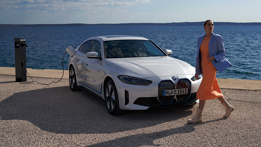 Vista anteriore a tre quarti della BMW i4 eDrive40 G26 2021 presso stazione di ricarica Una donna davanti alla BMW i4 eDrive40 G26 2021 Mineral White vista anteriore a tre quarti vicino alla colonnina di ricarica sul lago