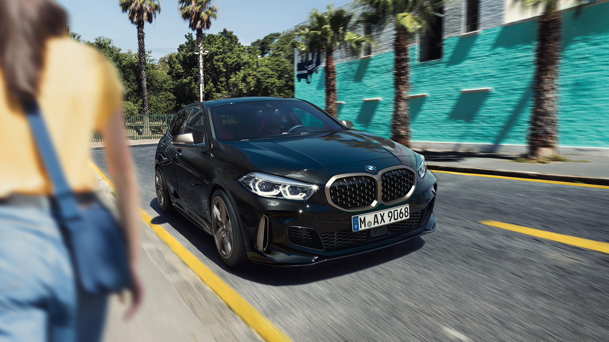 BMW M135i xDrive, vista anteriore a tre quarti. F40 BMW M135i xDrive in Sapphire Black metallizzato, vista anteriore a tre quarti con donna in primo piano. F40