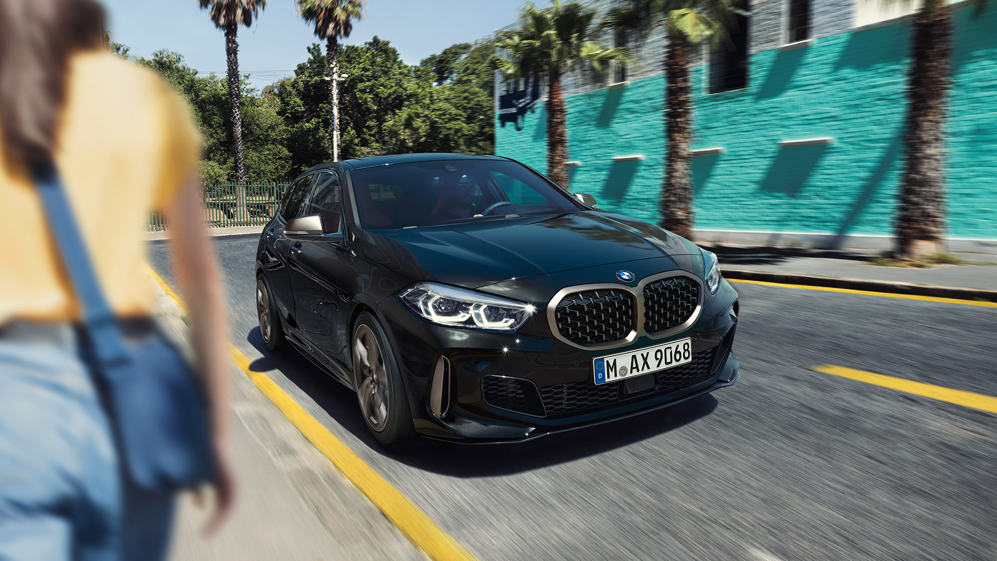 BMW M135i xDrive, vista anteriore a tre quarti. F40 BMW M135i xDrive in Sapphire Black metallizzato, vista anteriore a tre quarti con donna in primo piano. F40