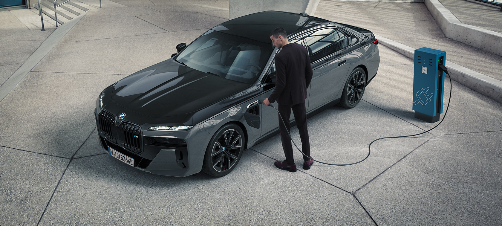 BMW M760e xDrive Berline G70 M Performance Pack BMW M760e xDrive Berline G70 M Performance Pack BMW teinte bi-ton BMW Individual Saphirschwarz metallic BMW Individual Dravitgrau metallic vue de trois quarts avant homme avec câble de recharge à côté d’une borne de recharge