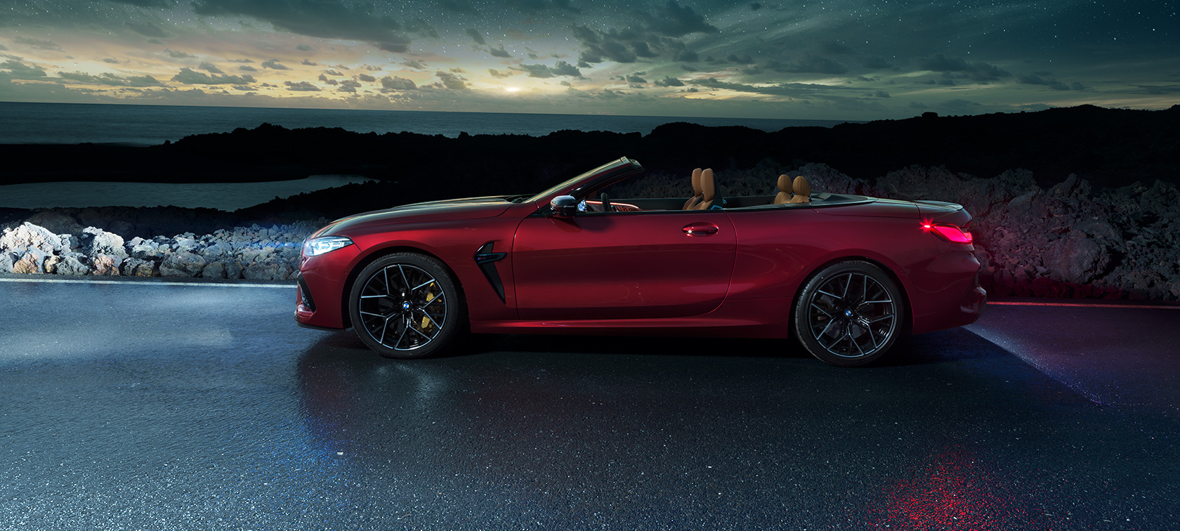 BMW M8 Competition Cabriolet, vista laterale. F91 BMW M8 Competition Cabriolet in Motegi Red metallizzato con capote aperta, vista laterale, in sosta su una strada costiera sotto il cielo stellato. F91