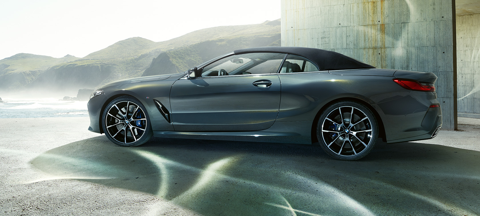 BMW M850i xDrive Cabriolet, vue de profil. G14 BMW M850i xDrive Cabriolet en Dravit Grau metallic avec capote fermée, vue de profil devant des collines. G14