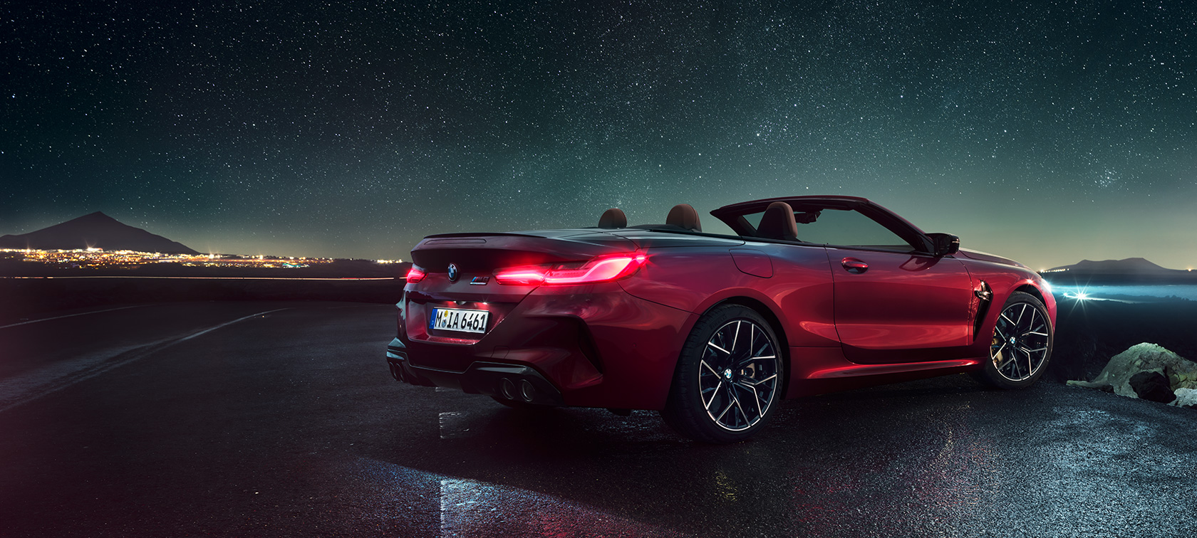 BMW M8 M Competition Cabriolet, vista posteriore di tre quarti. F91 BMW M8 Competition Cabriolet in Motegi Red metallizzato con capote aperta, vista posteriore di tre quarti, in sosta sotto il cielo stellato. F91