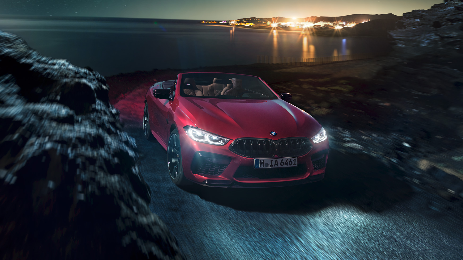 BMW M8 M Competition Cabriolet, vista laterale di tre quarti. F91 BMW M8 Competition Cabriolet in Motegi Red metallizzato con capote aperta, vista frontale di tre quarti, in marcia di notte su strada costiera. F91