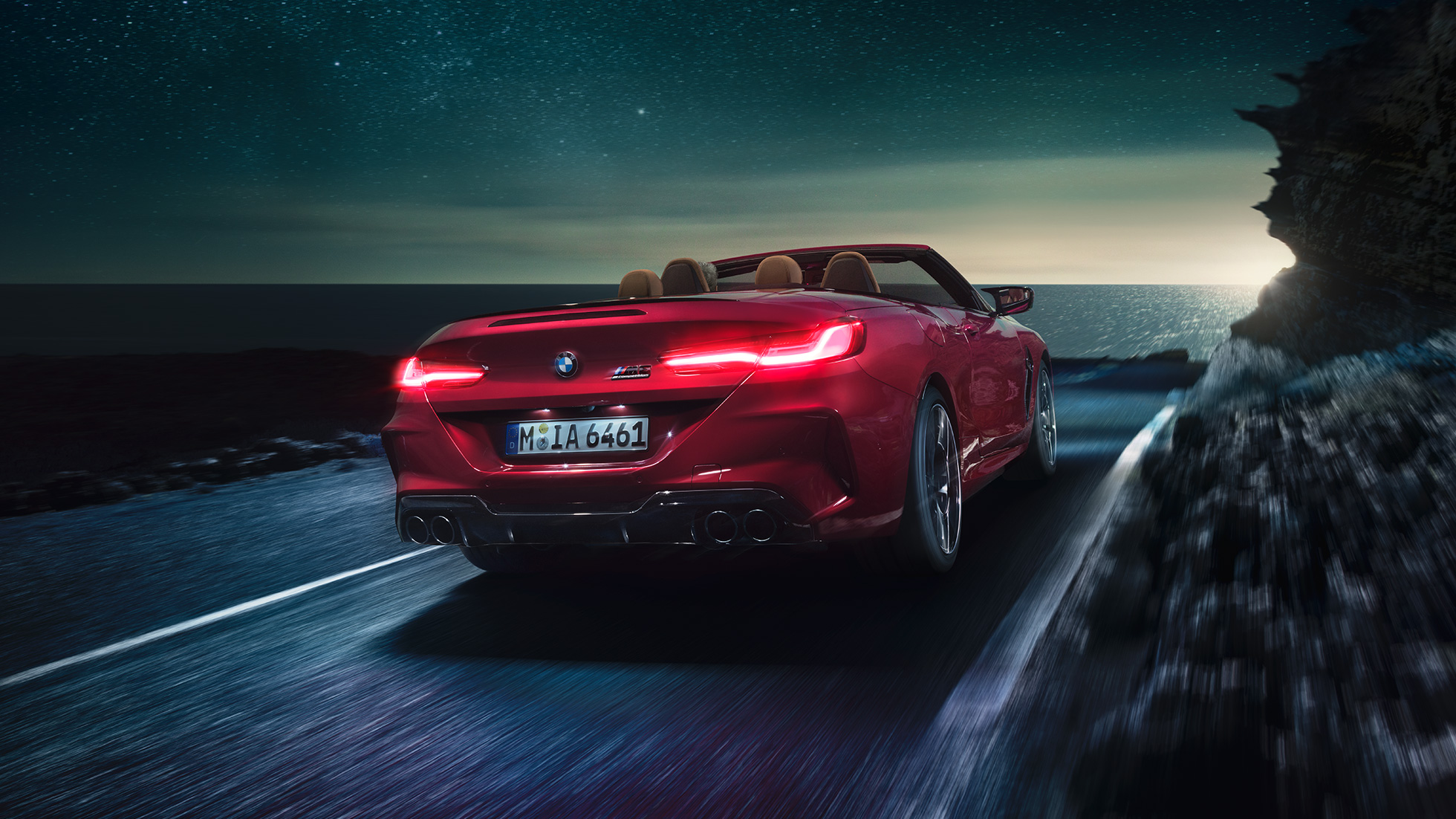BMW M8 M Competition Cabriolet, vista posteriore di tre quarti. F91 BMW M8 Competition Cabriolet in Motegi Red metallizzato con capote aperta, vista posteriore di tre quarti, in marcia di notte su strada costiera. F91