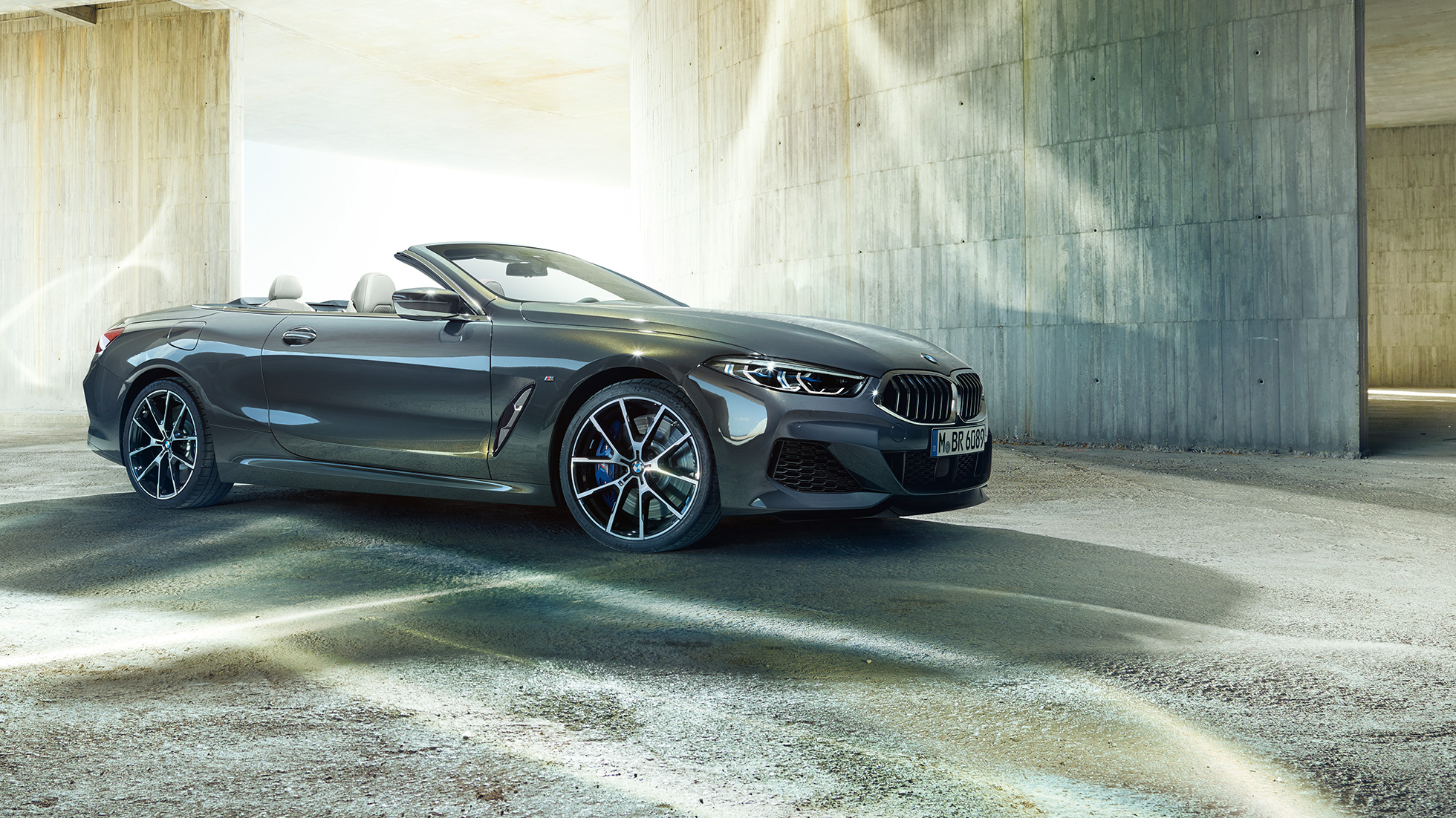 BMW M850i xDrive Cabriolet, vista laterale di tre quarti. G14 BMW M850i xDrive Cabriolet in Dravit Grey metallizzato con capote aperta, vista laterale di tre quarti, in sosta davanti a un edificio in calcestruzzo. G14