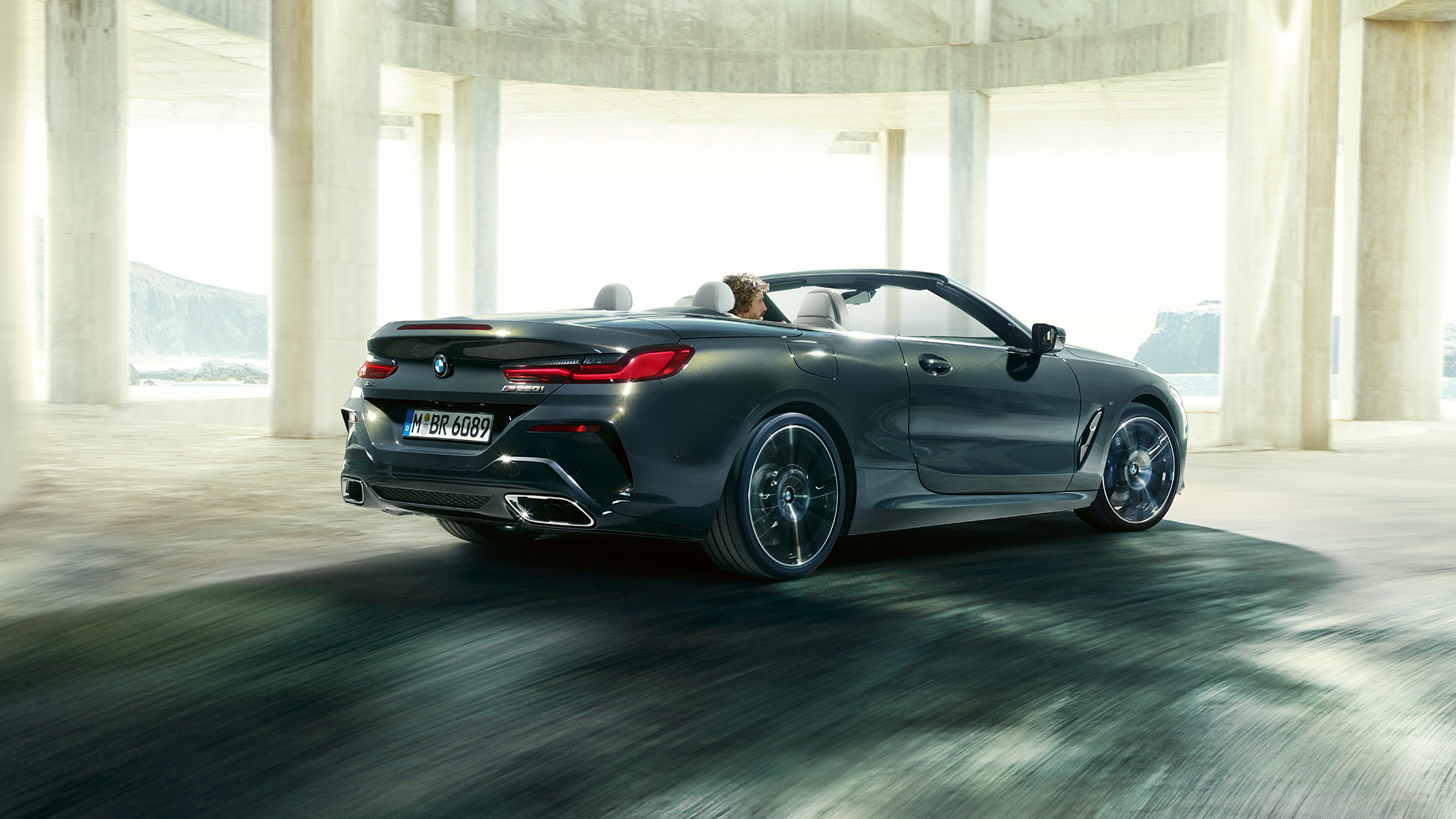 BMW M850i xDrive Cabrio, vista posteriore di tre quarti. G14 BMW M850i xDrive Cabrio in Dravit Grey metallizzato con capote aperta, vista posteriore di tre quarti, in marcia attraverso un edificio in calcestruzzo. G14