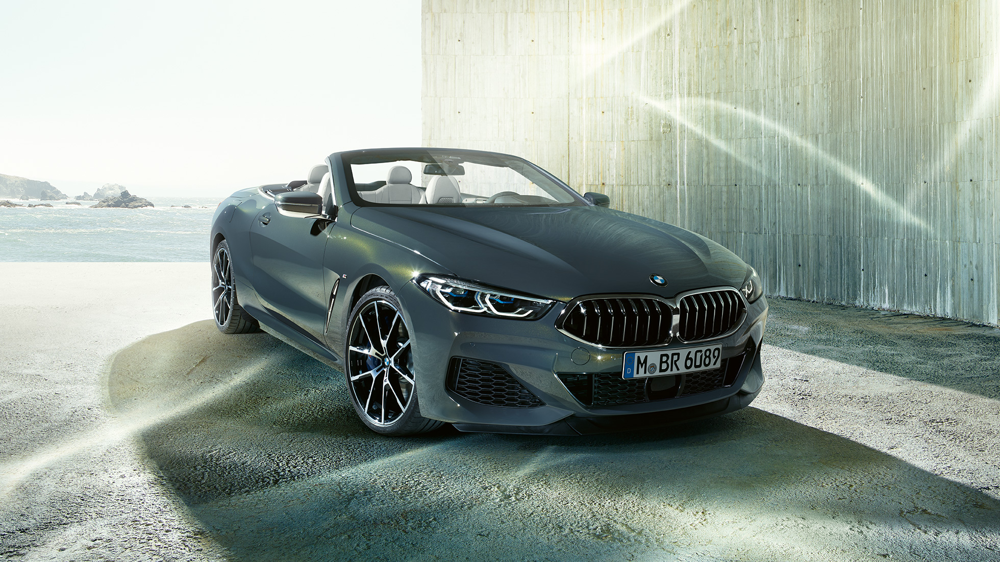BMW M850i xDrive Cabriolet, vista frontale di tre quarti. G14 BMW M850i xDrive Cabriolet in Dravit Grey metallizzato con capote aperta, vista frontale di tre quarti, in sosta davanti alla costa. G14