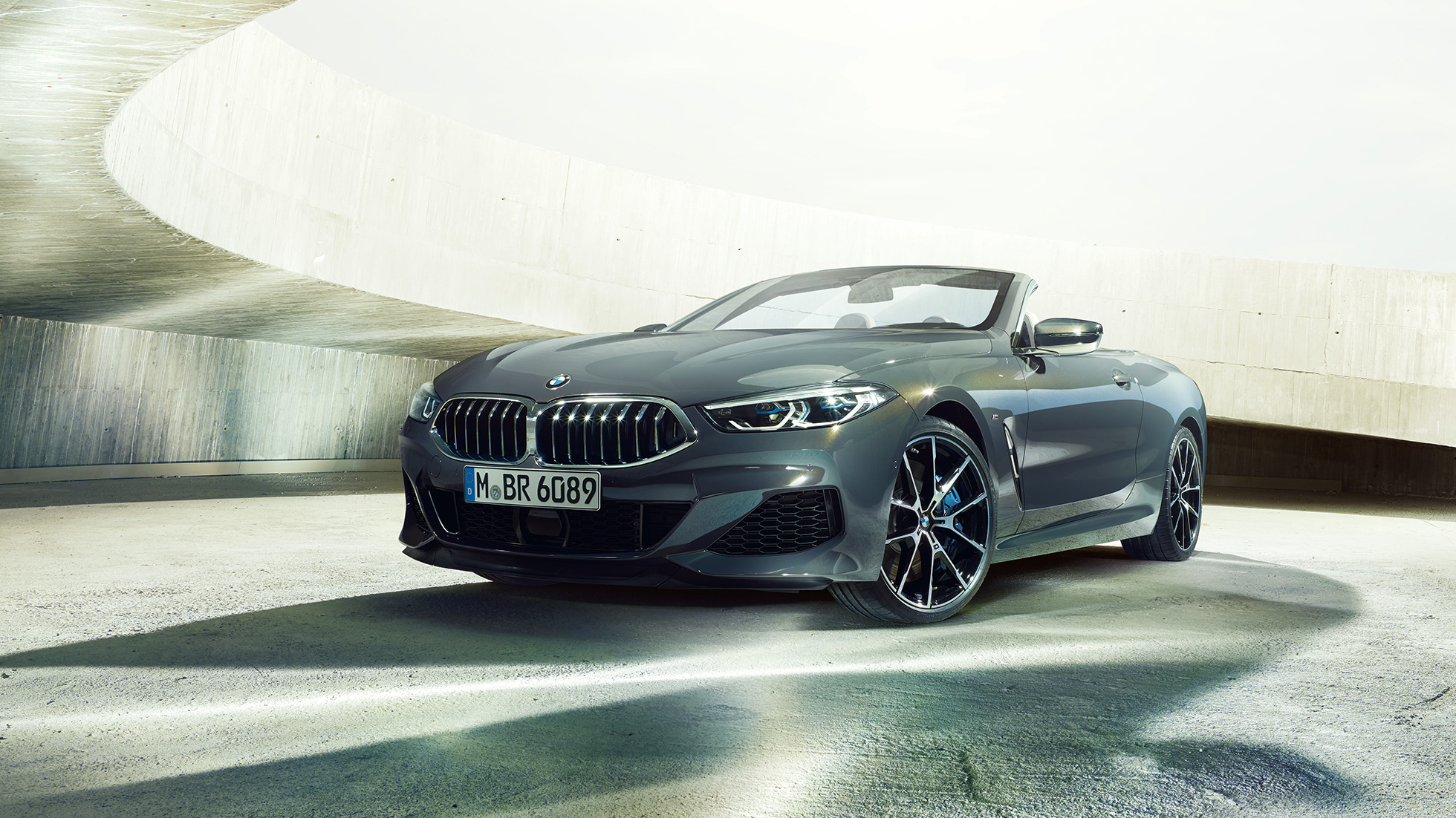 BMW M850i xDrive Cabriolet, vista frontale di tre quarti. G14 BMW M850i xDrive Cabriolet in Dravit Grey metallizzato con capote aperta, vista frontale di tre quarti, in sosta in un edificio in calcestruzzo. G14