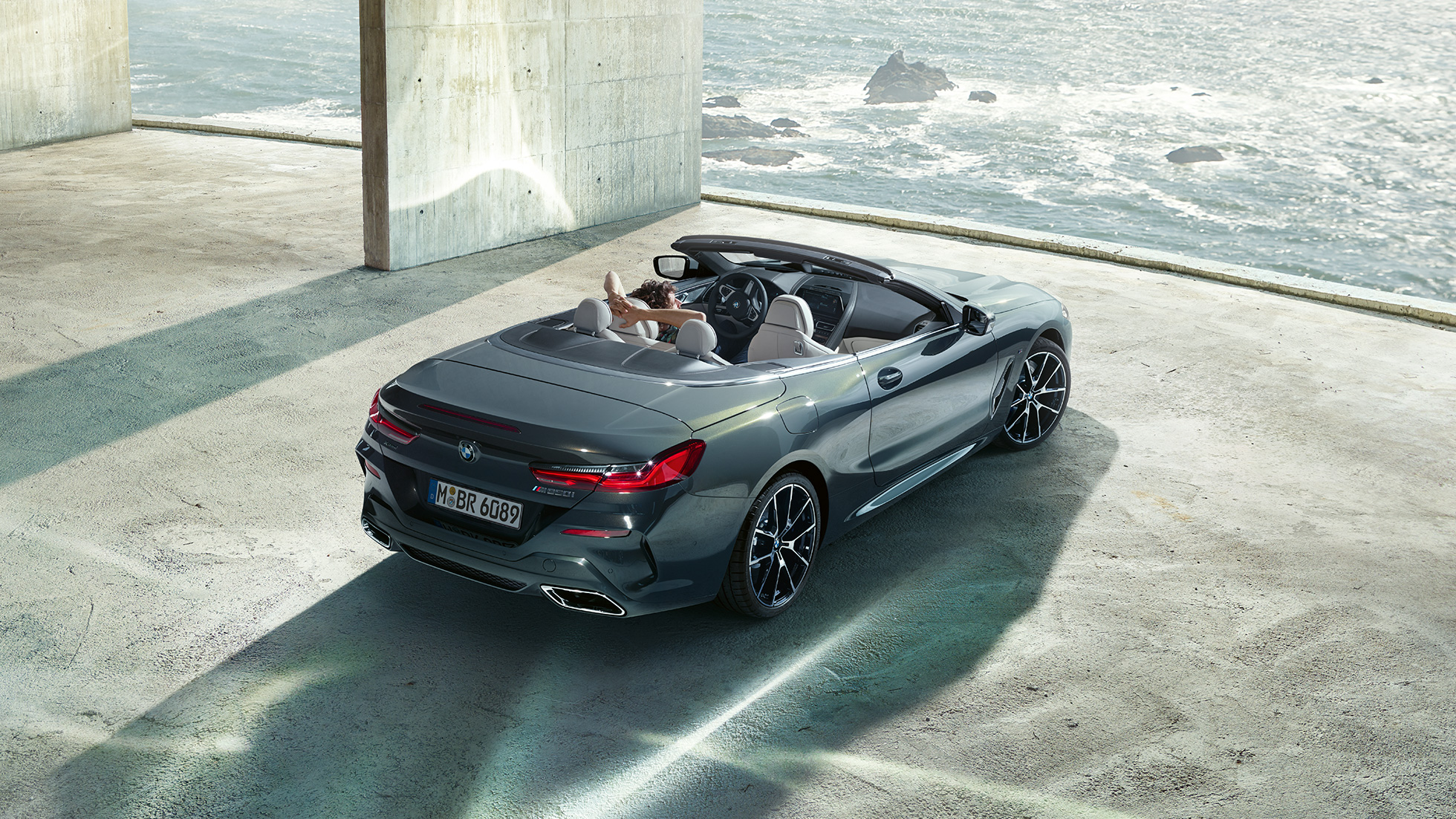 BMW M850i xDrive Cabriolet, prospettiva a volo d’uccello. G14 BMW M850i xDrive Cabriolet in Dravit Grey metallizzato con capote aperta, prospettiva a volo d’uccello, parcheggiata in edificio di calcestruzzo davanti all’oceano. G14