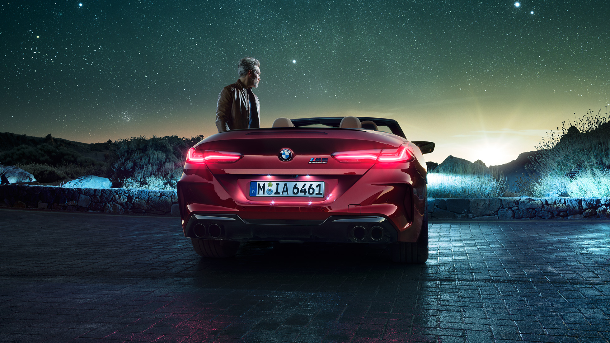 BMW M8 Competition Cabriolet, vista posteriore. F91 BMW M8 Competition Cabriolet in Motegi Red metallizzato con capote aperta, vista posteriore, in sosta con uomo sotto il cielo stellato. F91