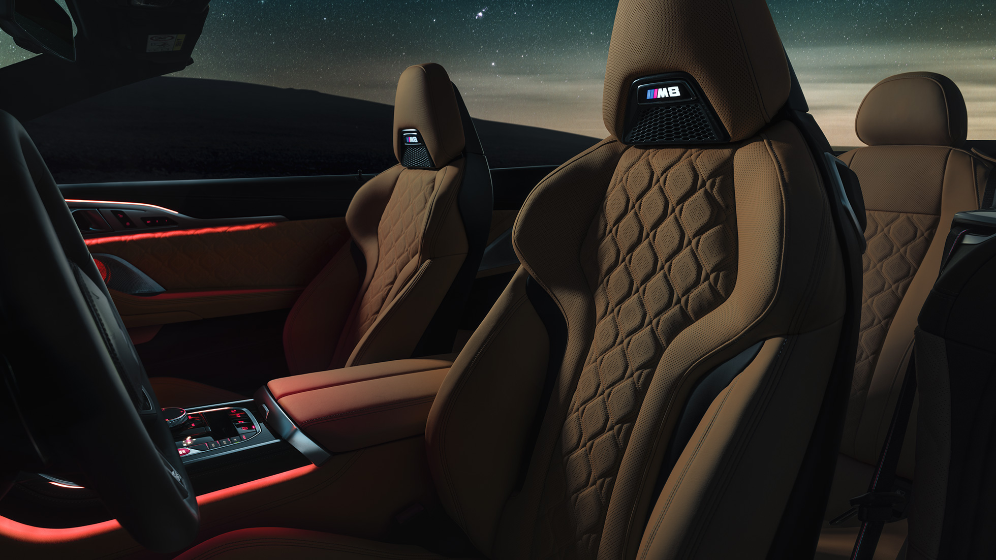 Sedili sportivi M in pelle Merino. F91 BMW M8 Competition Cabriolet con capote aperta sotto il cielo stellato, abitacolo con sedili sportivi M in pelle Merino e luce ambiente rossa. F91