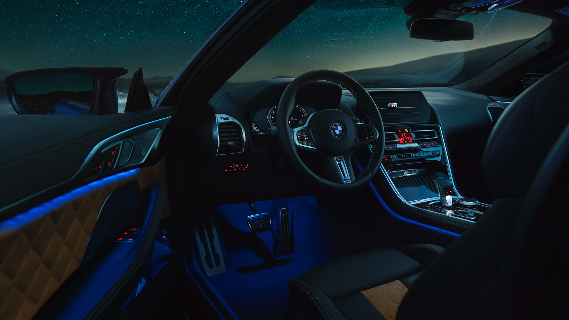 Interni BMW M8 Competition Coupé. F92 BMW M8 Competition Coupé, interni di notte con volante in pelle M e luce ambiente blu con porta del guidatore aperta. F92