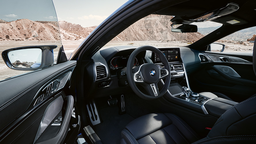 BMW M850i xDrive Coupé G15 LCI Facelift 2022 Cockpit orientato al guidatore con volante in pelle M e portiera del guidatore aperta