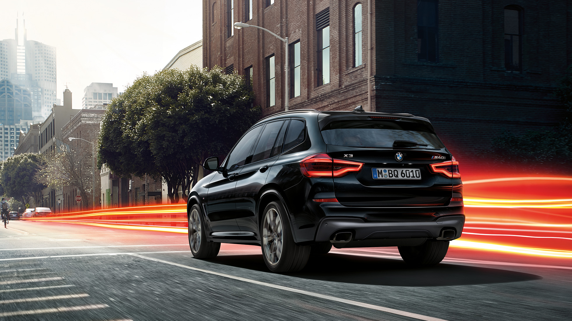 BMW X3 M40i, vista posteriore a tre quarti. BMW X3 M40i in Sapphire Black metallizzato, in movimento, esterni, vista posteriore a tre quarti su sfondo urbano con strisce di luce.