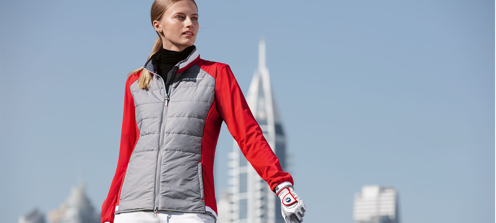 BMW Golfsport Jacke Damen. Auf dem Bild präsentiert eine junge Dame hochwertige Produkte aus der BMW Golfsport Collection vor einer Grossstadt-Skyline.