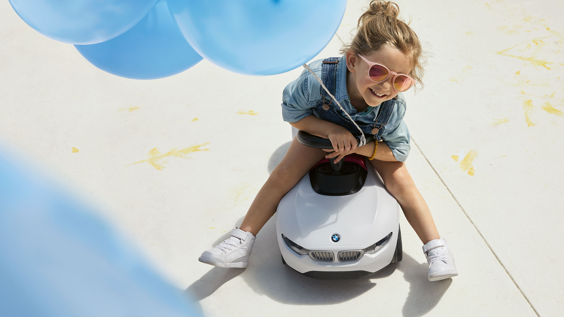 BMW Kids Collection. Ein Mädchen mit rosa Sonnenbrille und einem blauen Ballon in der Hand sitzt fröhlich auf einem BMW Babyracer.