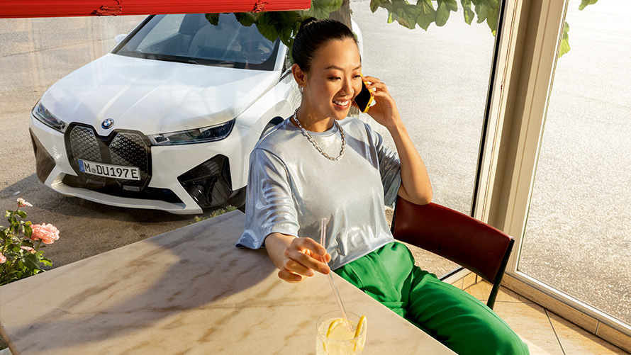 Condizioni di funzionamento di Proactive Care, check up & manutenzione Condizioni preliminari per Proactive Care, check up & manutenzione Donna seduta a tavola con smartphone in mane e con vettura BMW sullo sfondo