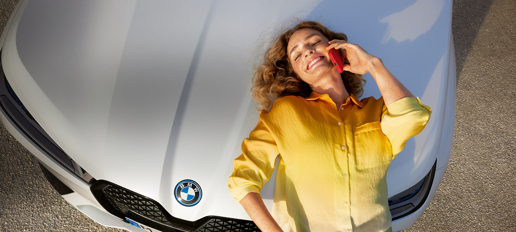 Relax We Care Proactive Care, Check-up & Wartung Proactive Care, Check-up & Wartung Relax We Care Frau mit gelbem Hemd und Smartphone in der Hand anlehnend auf weisser BMW Motorhaube