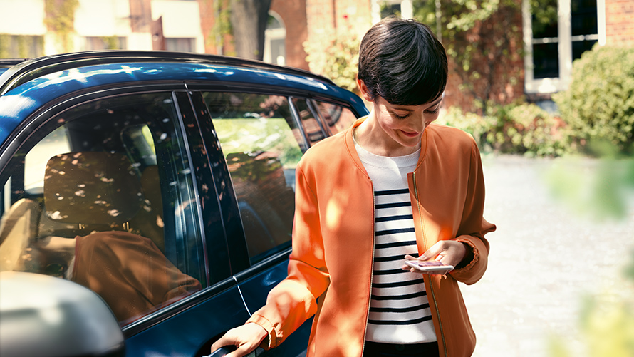 Im Leasing integrierte BMW Fahrzeugversicherung: Monatsprämie entspricht Modellbezeichnung. Lachende Frau steht neben parkiertem Auto mit Handy in der Hand.