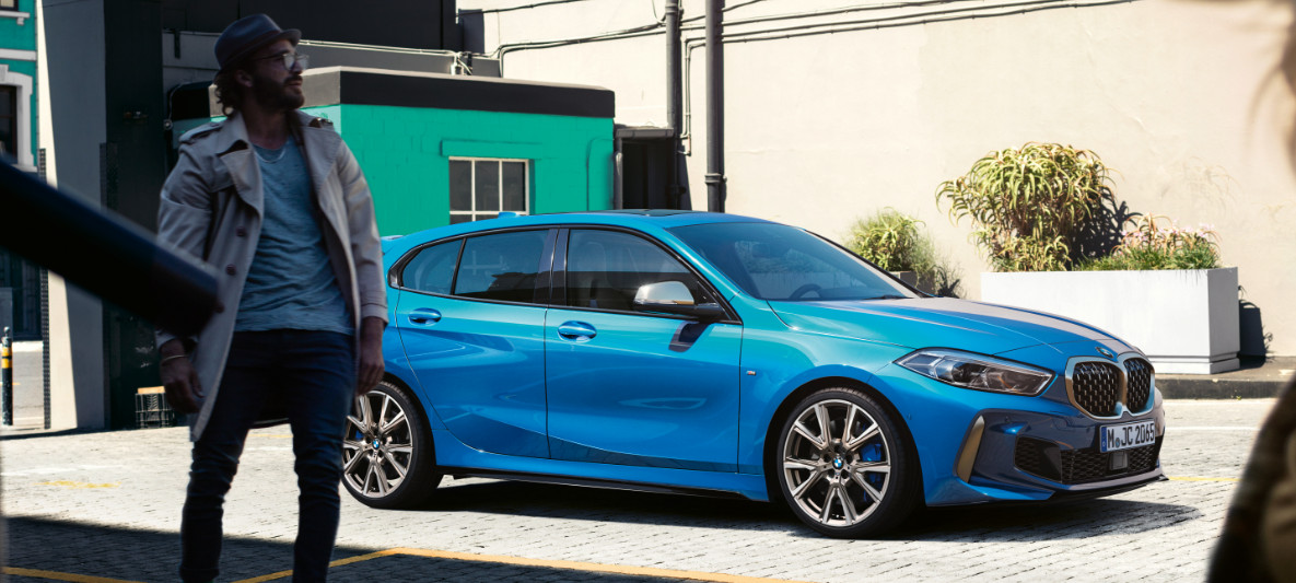 BMW 118i Essential ab CHF 199.–/Monat. BMW 118i Essential Edition parkiert auf Pflastersteinplatz, Mann im Vordergrund