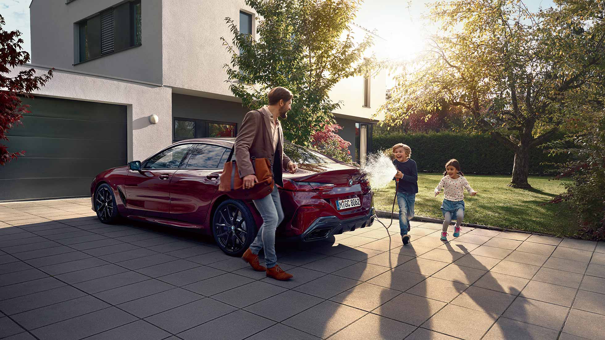 Pacchetto All In BMW, veicolo desiderato Padre e figlio nel parcheggio, davanti a una BMW ferma. Il figlio spruzza il padre con un tubo da giardino.