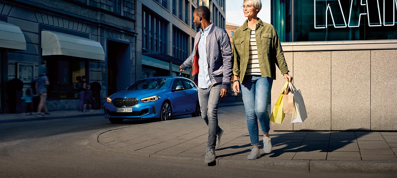 Pacchetto All In BMW. Il leasing comprende assicurazione, assistenza e garanzia commerciale. Donna e uomo attraversano la strada, l’uomo si gira a guardare una BMW Serie 1 blu.