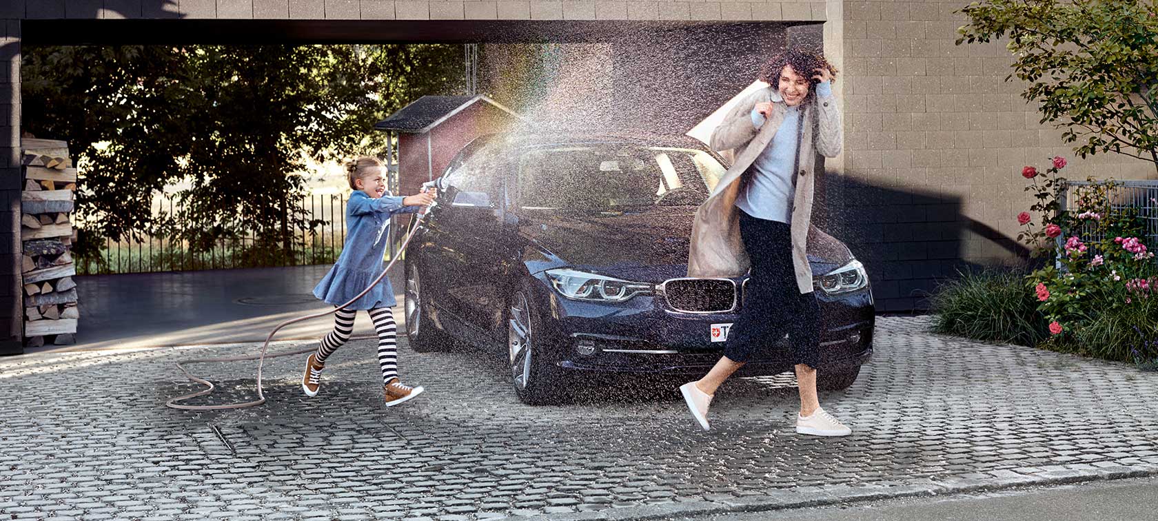 Offerta premium BMW: Occasioni di qualità con leasing allo 0,9% per la stipula di un’assicurazione auto e un’assicurazione rate BMW. Madre e figlia nel parcheggio, davanti a una BMW ferma. La figlia spruzza la mamma con un tubo da giardino.